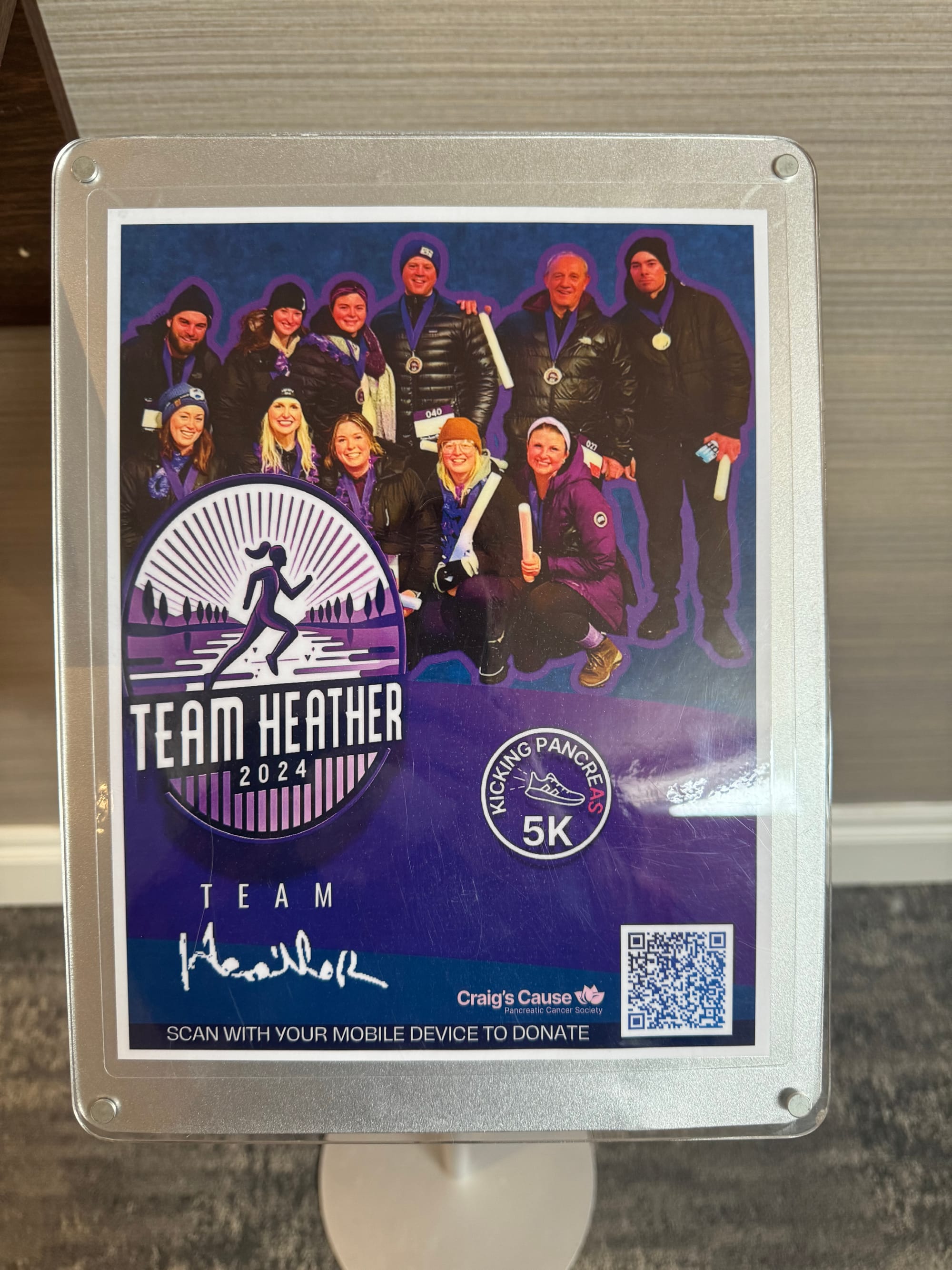 A custom designed purple poster displaying a photo of Team Heather from 2024, with a QR code encouraging funeral visitors to donate directly to Craig’s Cause on behalf of Heather.