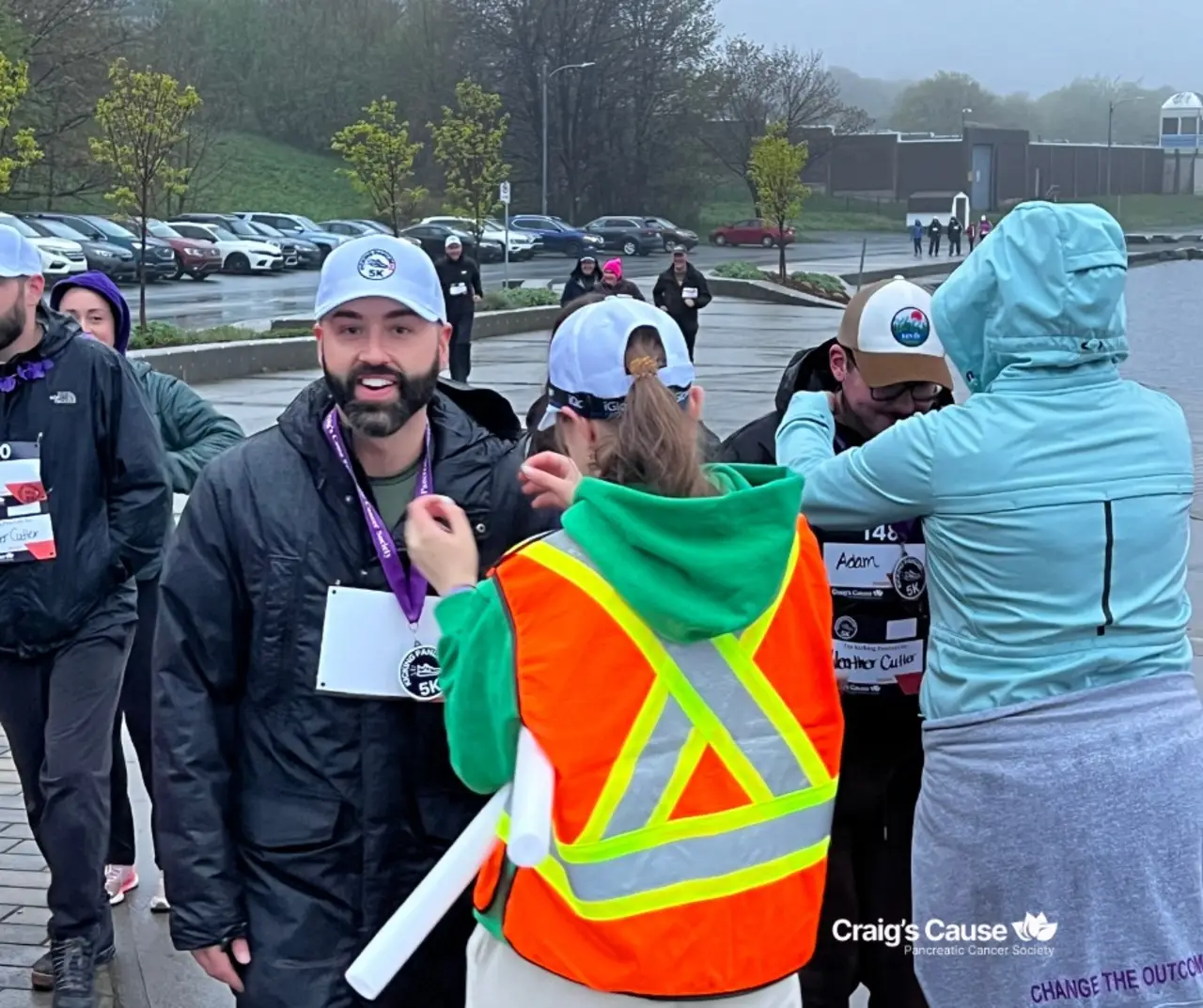 Participants at Craig’s Cause Pancreatic Cancer Society’s 5K Run/Walk being awarded medals and interacting with volunteers, on a rainy day with parked cars and other participants in the background.