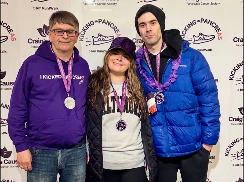 Three participants standing in front of a banner for Craig's Cause Pancreatic Cancer Society's 5K Run/Walk event, smiling and wearing medals and event gear, showing support for pancreatic cancer awareness."