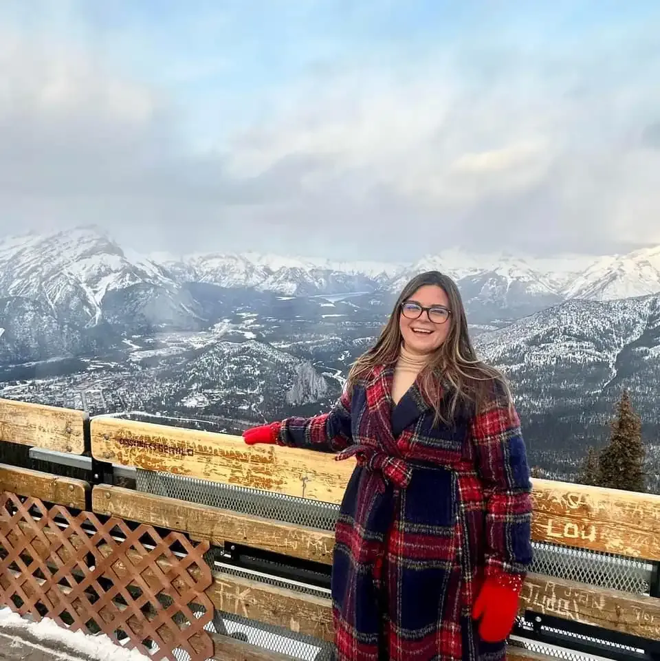 A person wearing a plaid coat and red gloves stands smiling on a snowy mountain viewpoint with a vast landscape of snowy mountains and valleys in the background, enjoying the moment.