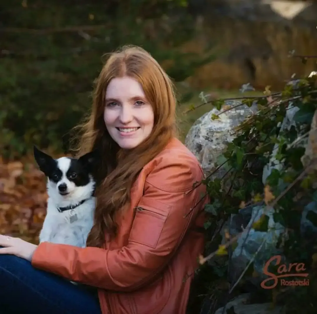 A woman with long red hair wearing an orange jacket sits outdoors with a small black and white dog on her lap. The background includes greenery and rocks.