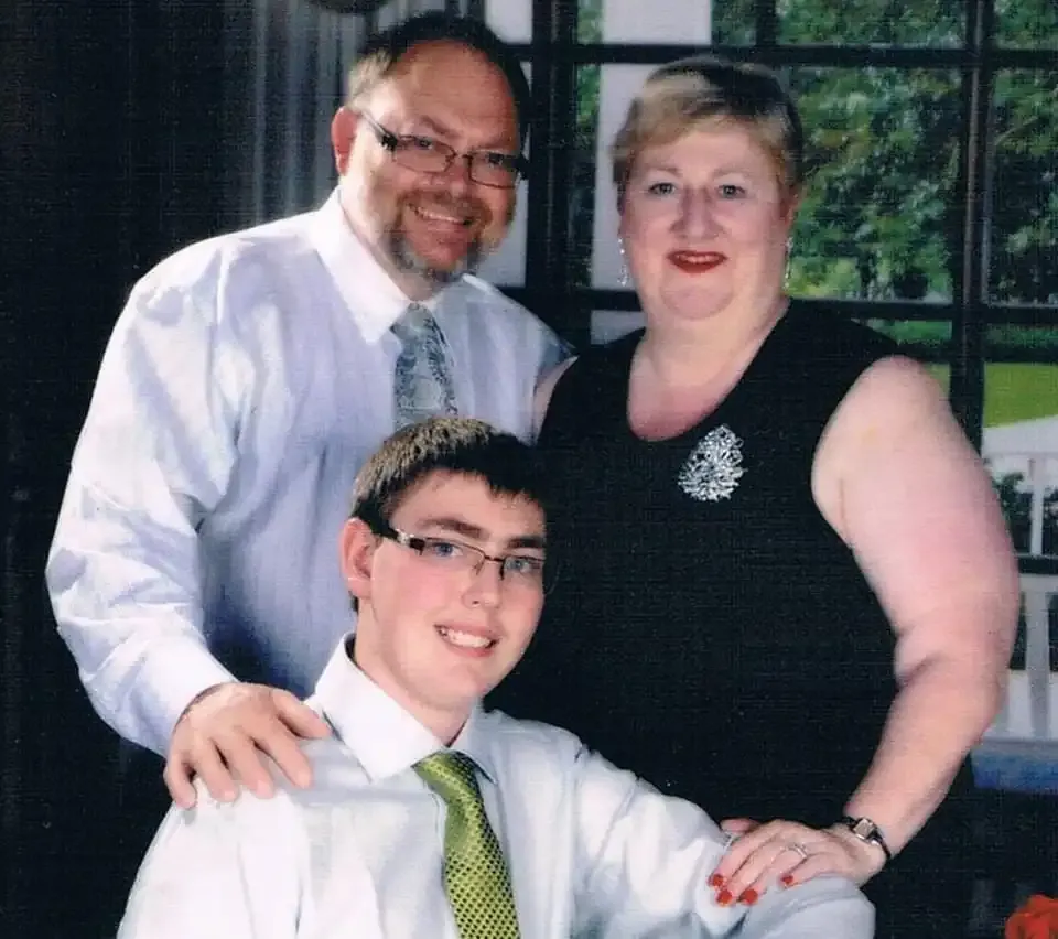 A smiling family of three posing indoors, with the son seated and parents standing behind him in formal attire, radiates warmth. Their strength shines through as lifelong pancreatic cancer advocates and caregivers.