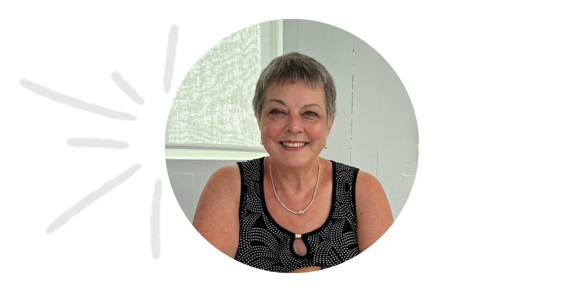 A woman with short gray hair smiles at the camera, wearing a black patterned sleeveless top and a silver necklace. Seated indoors against a light-colored background, she represents Team Heather in the "About Us" section.