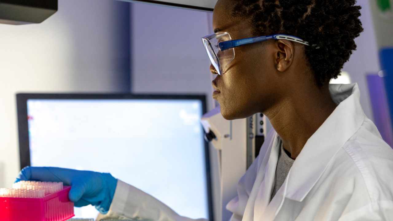 A cancer researcher in a lab coat, wearing blue gloves, works in a Canadian lab. A monitor displays research funding data.