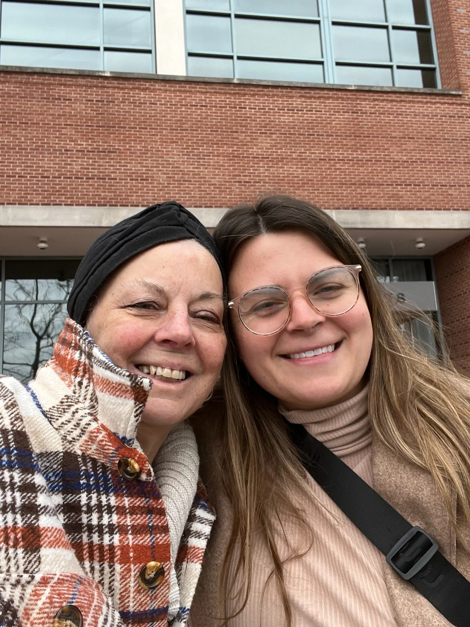Two women in winter coats smile together in a selfie outside a brick building, wearing plaid and beige coats on a cloudy day.