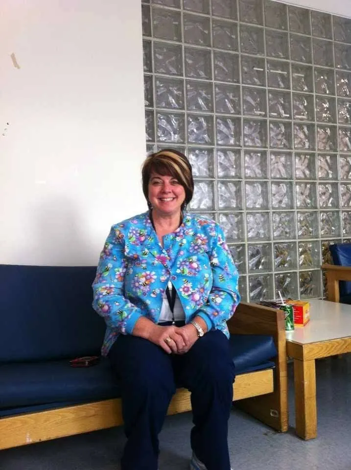 Woman in blue floral print scrub jacket sitting on a bench with glass block wall background in hospital break area.
