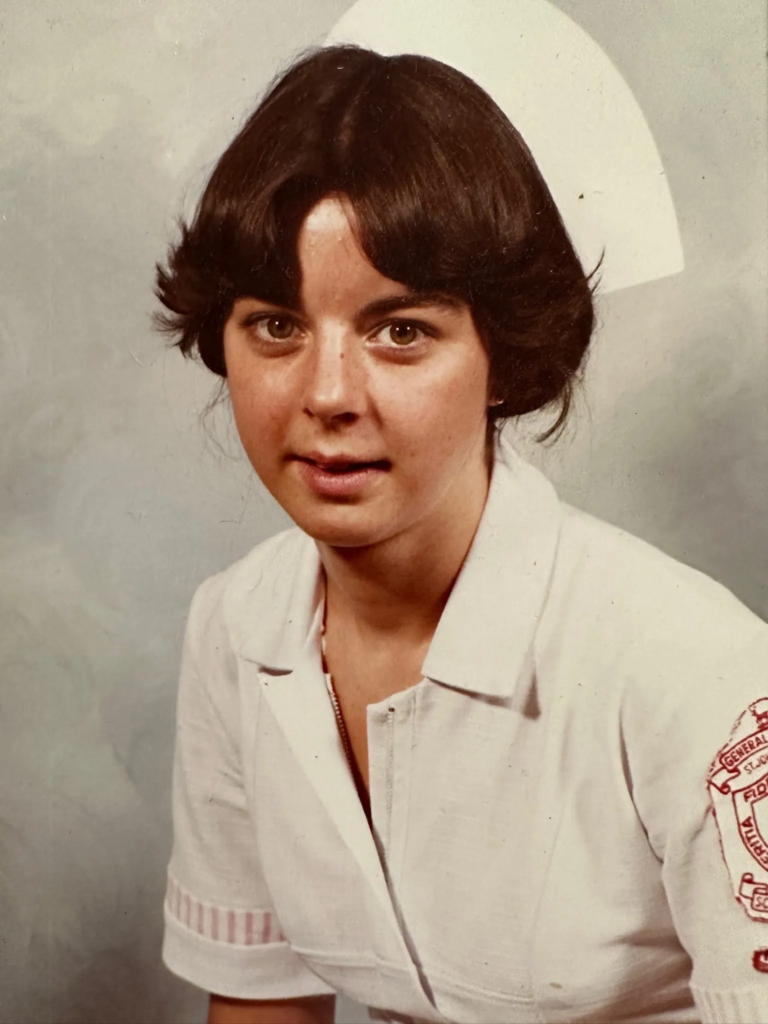 Portrait of a young woman in traditional white nursing uniform and cap against a neutral background, early 1980s graduation photo.