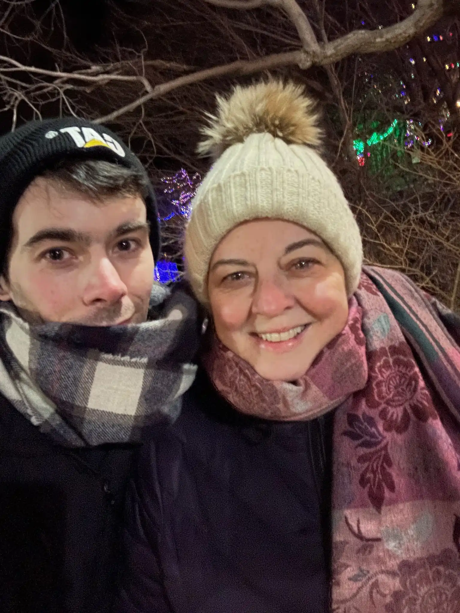 Nighttime winter selfie of mother and son wearing warm hats and scarves, smiling together with colorful holiday lights glowing in background.