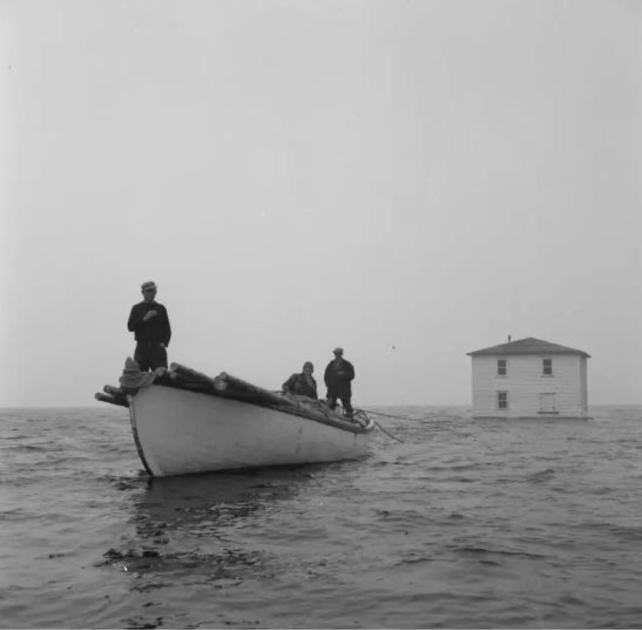 Black-and-white photograph of men in an open boat towing a two-storey house across the water, from the resettlement years when outport families moved their homes by sea.