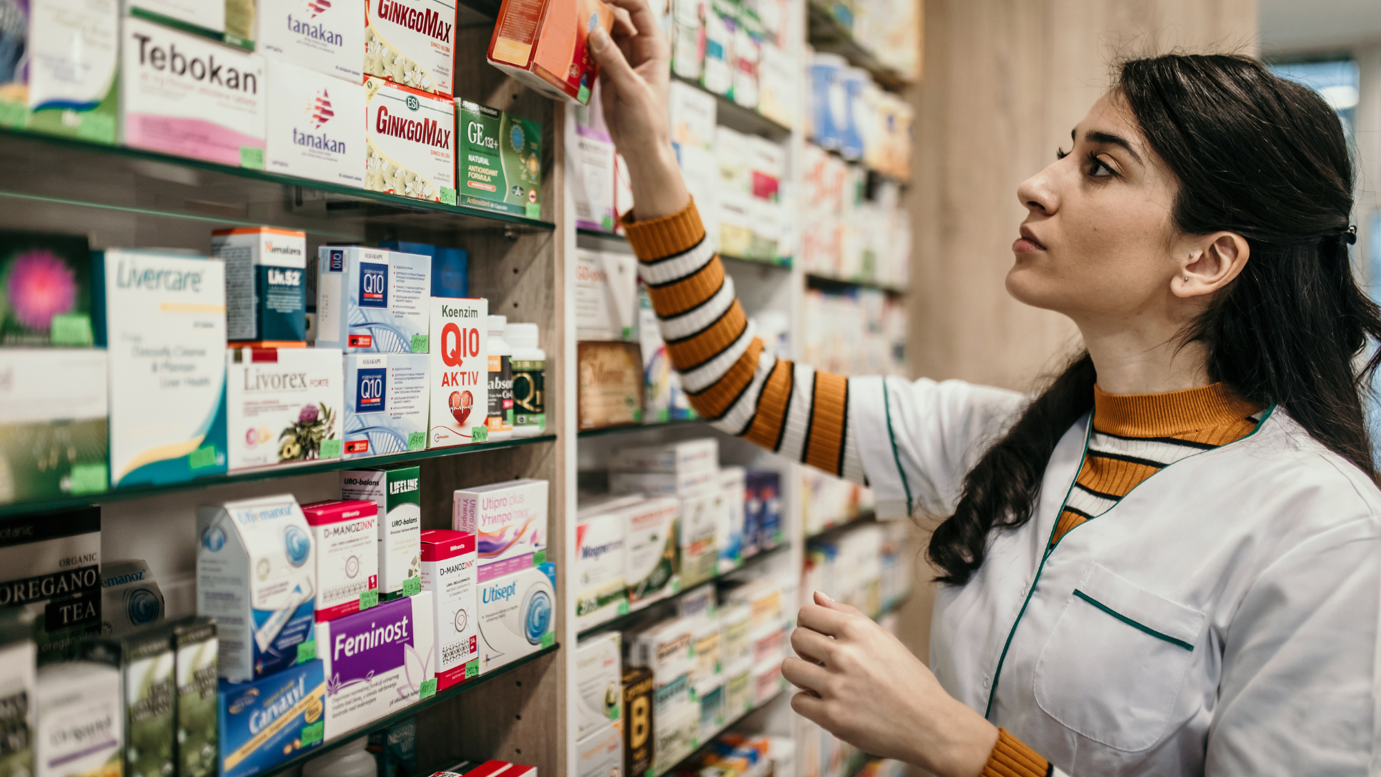 A pharmacist in a white coat reaching for a box of medication on a pharmacy shelf.