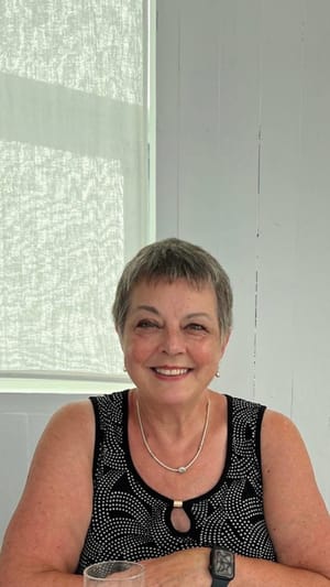 Heather Cutler sits smiling at a white table, wearing a black patterned top and silver jewelry, beneath a painted purple arc.