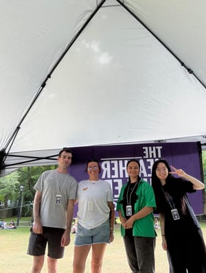 Four volunteers from The Heather Cutler Foundation smile under a canopy at Pride, standing in front of a purple banner.